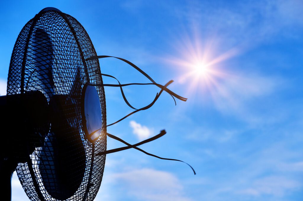 Ein laufender Ventilator, der vor einem blauen Himmel mit glühender Sonne steht und für Abkühlung sorgen soll.