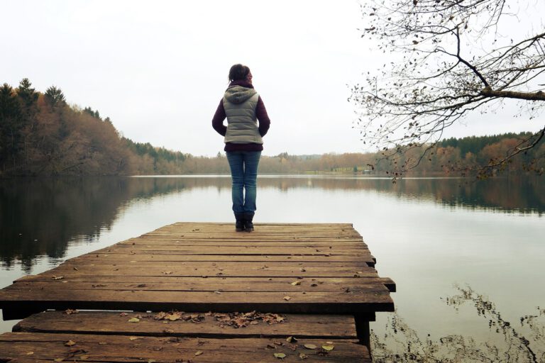 An einem düsteren Herbsttag steht eine Frau steht auf einem Steg und blickt nachdenklich auf einen ruhigen See.