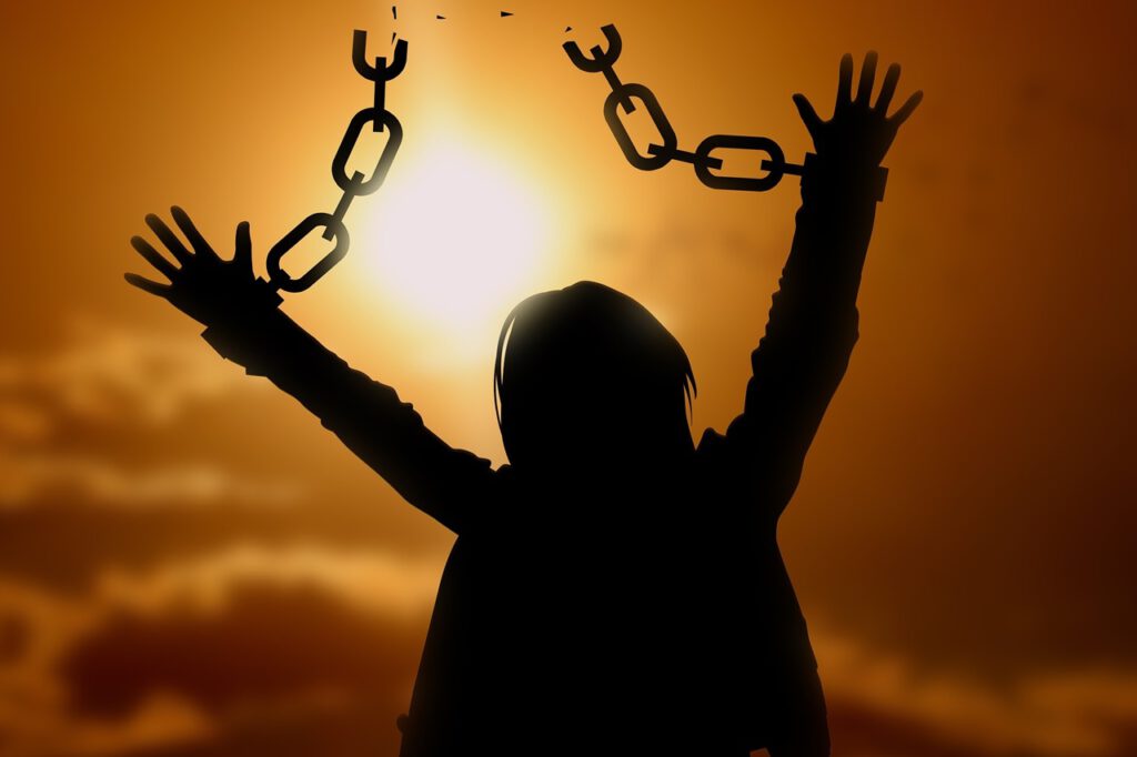 Eine Frau sieht in den Sonnenuntergang und feiert ihre Freiheit. Sie streckt ihre Arme in die Höhe, an denen Ketten befestigt waren.