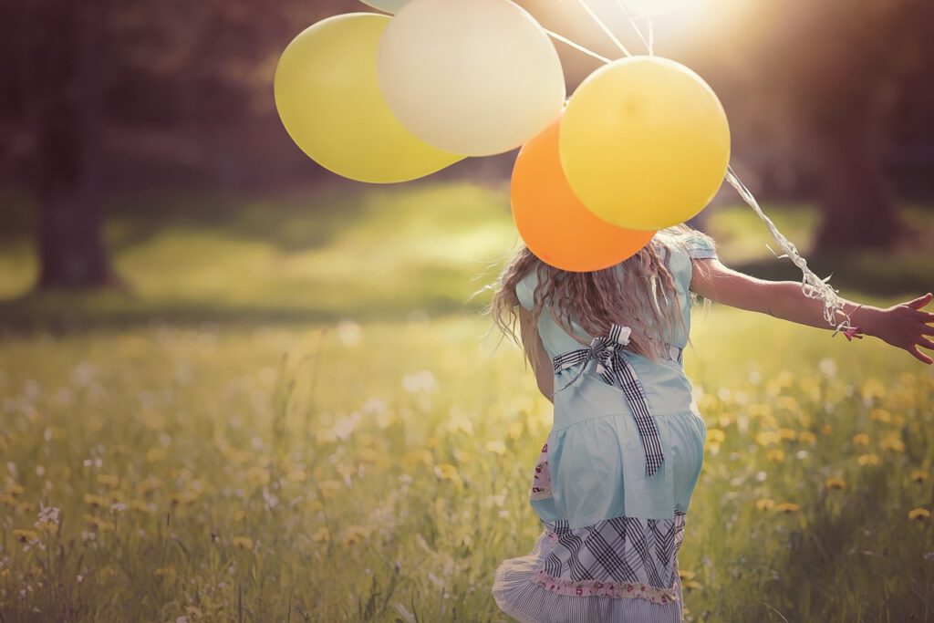 Ein Mädchen mit bunten Luftballons läuft fröhlich über eine Blumenwiese.