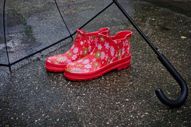 An einem Regentag stehen auf dem nassen Asphalt ein paar rote Gummistiefel neben einem Regenschirm.