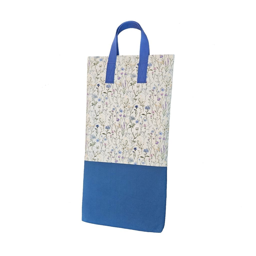 Eine stabile schmale, aber hohe Stofftasche. Der untere Teil der Tasche ist blau. Der obere Teil ist eine bunte Blumen-Optik auf hellgrauem Hintergrund. Die Henkel sind passend blau.