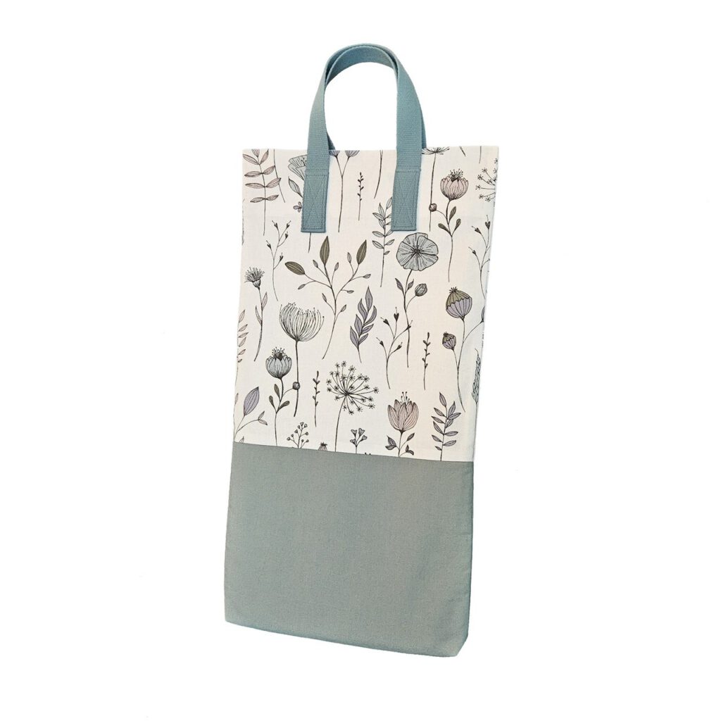 Eine stabile schmale, aber hohe Stofftasche. Der untere Teil der Tasche ist pastell-grün. Der obere Teil ist eine Blumen-Optik auf weißem Hintergrund. Die Henkel sind passend grün.