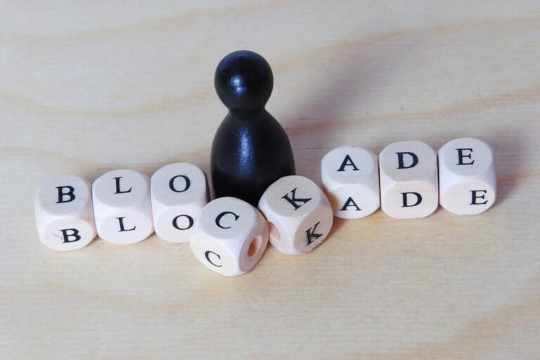 Schwarze Spielfigur steht auf hellem Holzuntergrund umgeben von weißen Würfeln mit den Buchstaben 'BLOCKADE'