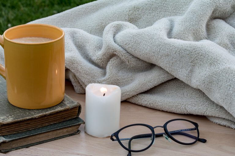 Becher mit Getränk auf zwei Büchern neben brennender Kerze, Brille und weicher Decke auf Holztisch
