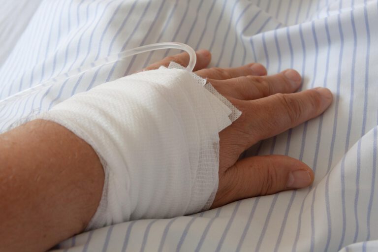 Eine verbundene Hand mit Infusionsschlauch auf gestreifter Bettdecke.