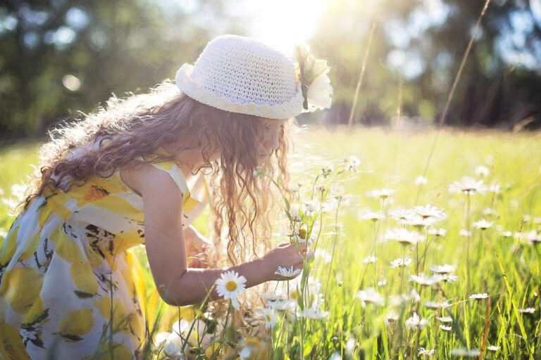 Eine junge Frau im Sommerkleid und Sommerhut steht in einem Feld voller Margeriten und pflückt unbeschwert Blumen.