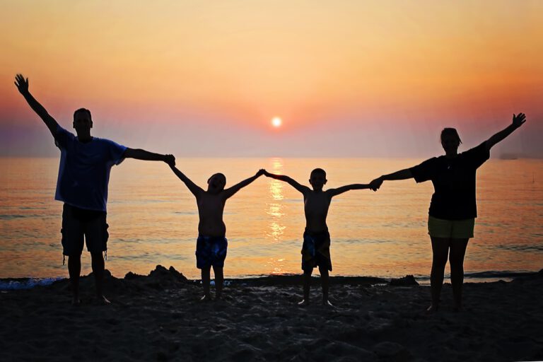 Eine vierköpfige Familie steht im warmen Gegenlicht des Sonnenuntergangs am Strand, hält sich an den Händen und blickt glücklich nach vorne.