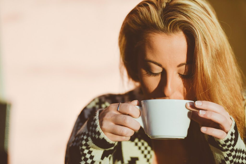 Eine erschöpfte Frau mit geschlossenen Augen trägt einen schwarz-weißen Pullover und hält eine weiße Kaffeetasse in den Händen.