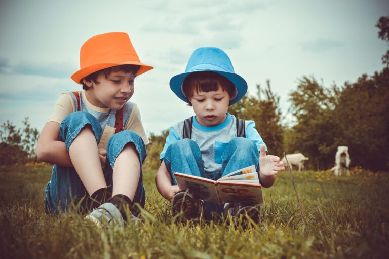 Zwei Jungs mit buntem Hut sitzen auf einer grünen Wiese und lesen zusammen ein Buch.